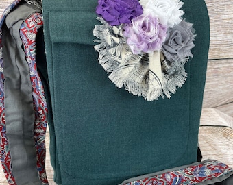 Puede incluir: Un bolso bandolera de color verde oscuro con un grupo decorativo de flores de tela en tonos morado, blanco, lavanda y gris. El bolso incluye una correa larga con un estampado de cachemir rojo, azul y blanco y un forro interior gris.