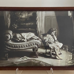 May include: A framed black and white print depicts two young children in a room. One child sleeps on a chaise lounge, while the other sits in a rocking chair. The print has a dark brown wooden frame.