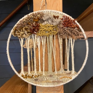 Puede incluir: Una pared circular hecha de hilo con un diseño tejido de árboles. Los árboles están hechos de hilo blanco y el fondo es una mezcla de hilo marrón, beige y amarillo.