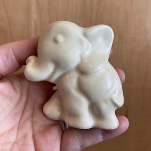 Könnte beinhalten: Eine kleine, weiße Keramik-Elefantenfigur. Der Elefant steht auf allen vieren und hat seinen Rüssel erhoben. Die Figur ist ein schlichtes, minimalistisches Design.