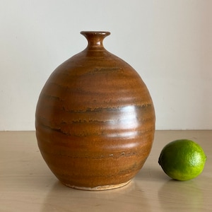 May include: A brown ceramic vase with a rounded body and a narrow neck. The vase has a streaky glaze and is sitting on a light wood surface.