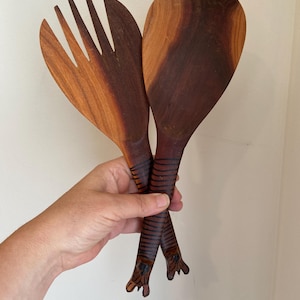 Puede incluir: Un juego de dos cucharas para ensalada de madera con mangos de rayas de tigre. Las cucharas son de madera oscura y tienen un acabado natural. Una cuchara tiene una cabeza en forma de cuchara, mientras que la otra tiene una cabeza en forma de tenedor.