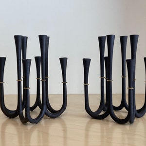 以下が含まれることがあります： モダンなデザインの黒い金属製のキャンドルホルダー2つ。各ホルダーには9つのキャンドル用スロットがあり、金色のアクセントが施されています。