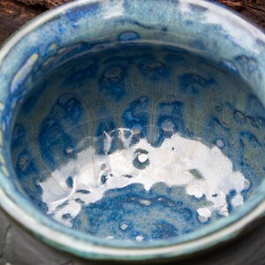 May include: A ceramic bowl featuring a glossy, reactive glaze in shades of deep cobalt blue, light sky blue, and hints of sage green. The interior surface displays a mottled, fluid pattern with a reflective finish and a smooth, rounded rim.