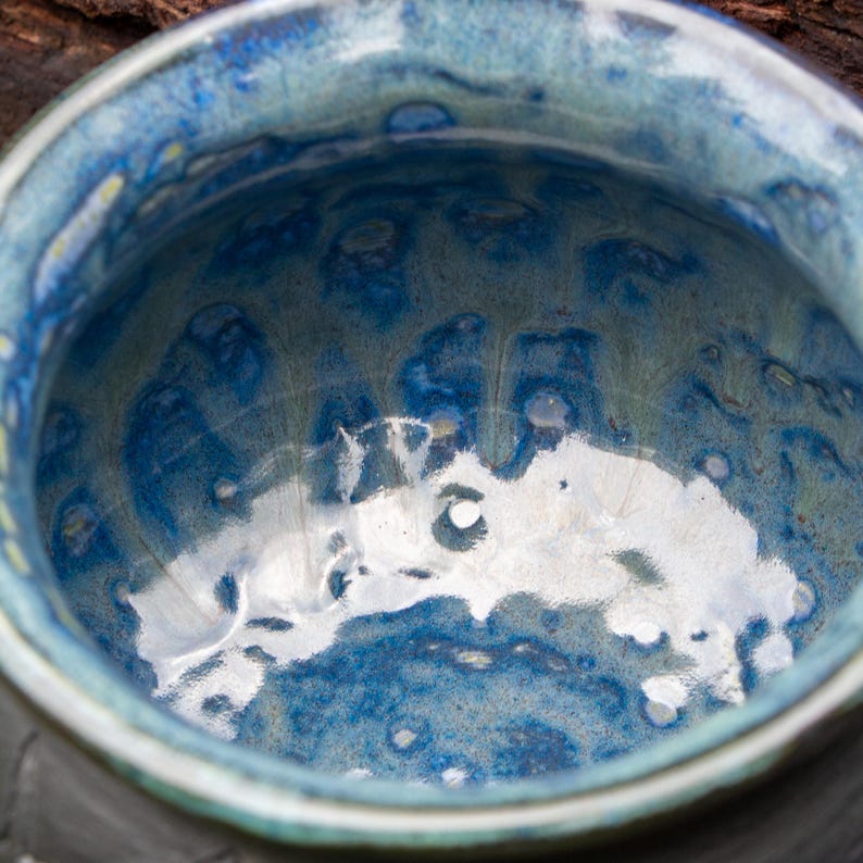 May include: A ceramic bowl featuring a glossy, reactive glaze in shades of deep cobalt blue, light sky blue, and hints of sage green. The interior surface displays a mottled, fluid pattern with a reflective finish and a smooth, rounded rim.