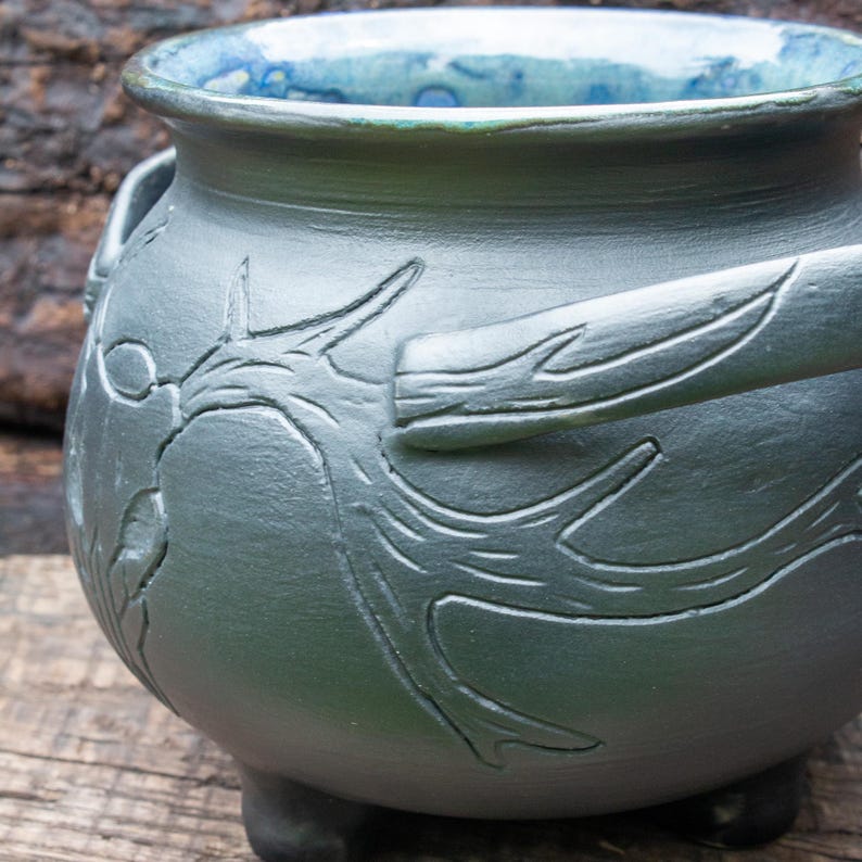 May include: A matte dark gray ceramic cauldron with a rounded body and three small feet. The exterior features a carved tree branch design wrapping around the sides. The interior of the vessel is glazed in a vibrant, glossy blue and green finish.