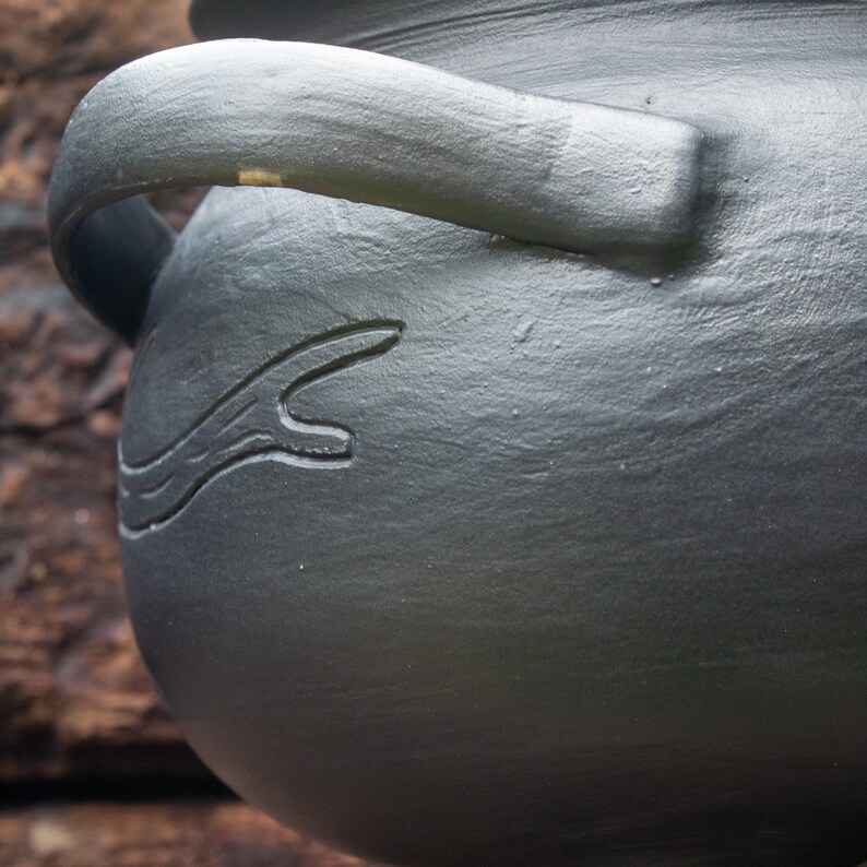 May include: A matte dark gray ceramic pot featuring a curved handle and a carved branch-like design on its rounded side. The surface has a textured, hand-crafted appearance with visible clay markings and a consistent dark gray finish throughout.