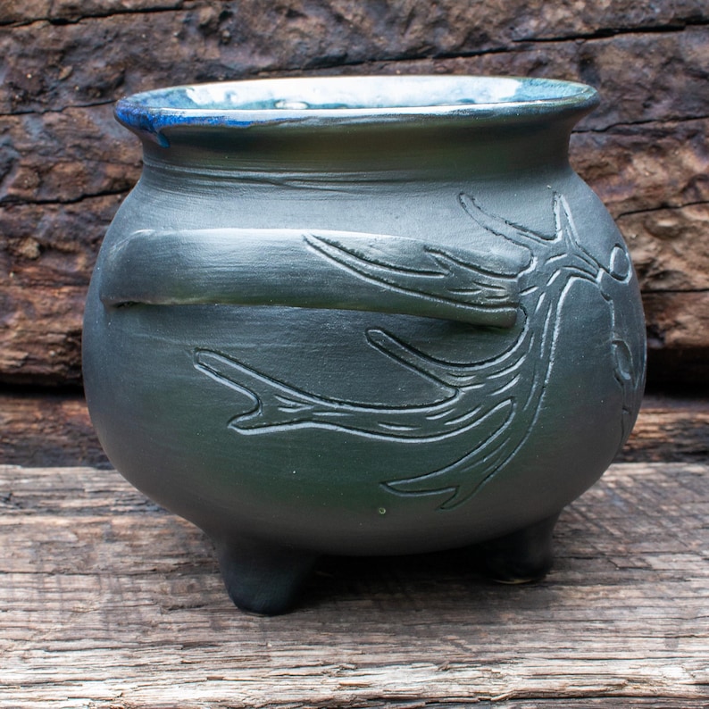 May include: A matte black ceramic cauldron with a rounded body, three short legs, and a wide rim featuring a blue interior glaze. The exterior surface is decorated with a raised, sculpted relief of a tree branch extending across the side.