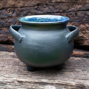 May include: A rounded ceramic cauldron with a matte charcoal black exterior and a glossy, mottled blue interior glaze. The vessel features two side handles, a wide circular opening, and three short, sturdy feet supporting the base.