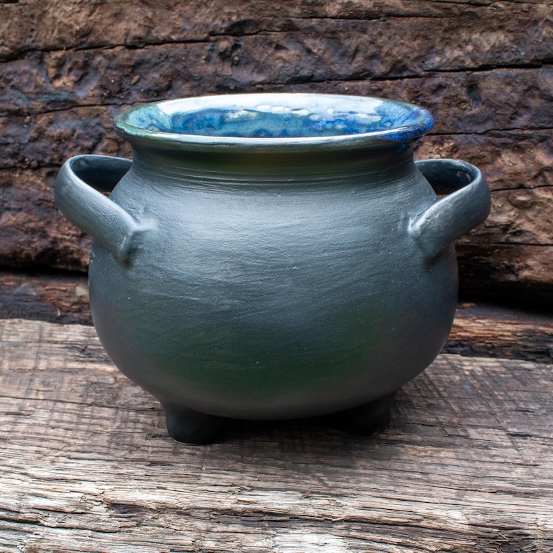 May include: A rounded ceramic cauldron with a matte charcoal black exterior and a glossy, mottled blue interior glaze. The vessel features two side handles, a wide circular opening, and three short, sturdy feet supporting the base.