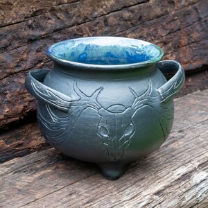 May include: A dark gray ceramic cauldron with two side handles and three small feet. The exterior features an embossed deer skull design with antlers extending toward the handles. The interior is glazed in a vibrant, mottled blue finish.