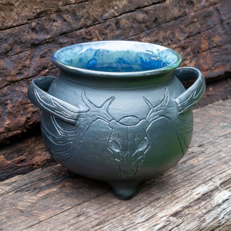 May include: A dark gray ceramic cauldron with two side handles and three small feet. The exterior features an embossed deer skull design with antlers extending toward the handles. The interior is glazed in a vibrant, mottled blue finish.