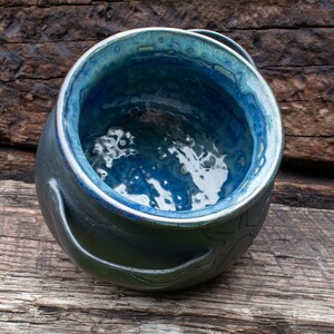 May include: A round ceramic pot featuring a deep, glossy blue interior glaze with a textured, mottled finish. The exterior is a dark, matte charcoal color with a smooth surface and a curved handle extending from the side of the vessel.