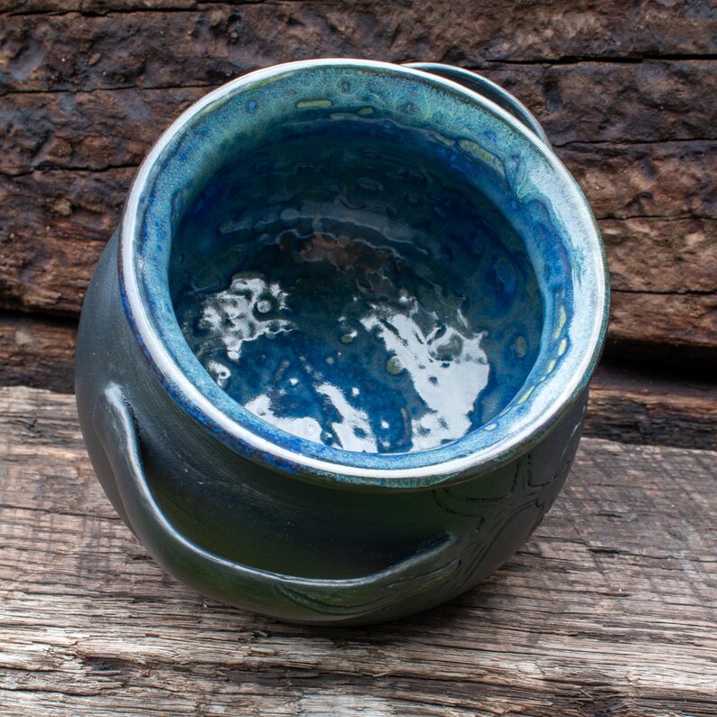 May include: A round ceramic pot featuring a deep, glossy blue interior glaze with a textured, mottled finish. The exterior is a dark, matte charcoal color with a smooth surface and a curved handle extending from the side of the vessel.