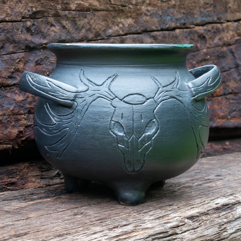 May include: A matte black ceramic cauldron featuring a carved deer skull design on the front. The vessel has a rounded body, a wide rim, two side handles, and three short feet. The intricate carving extends across the surface of the pot.