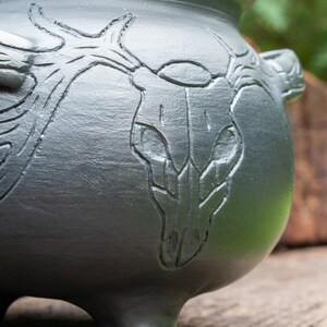 May include: A matte black ceramic cauldron featuring a deeply embossed design of a deer skull with large, sprawling antlers. The vessel has a rounded body, a wide opening at the top, and integrated side handles, with a smooth, dark finish.