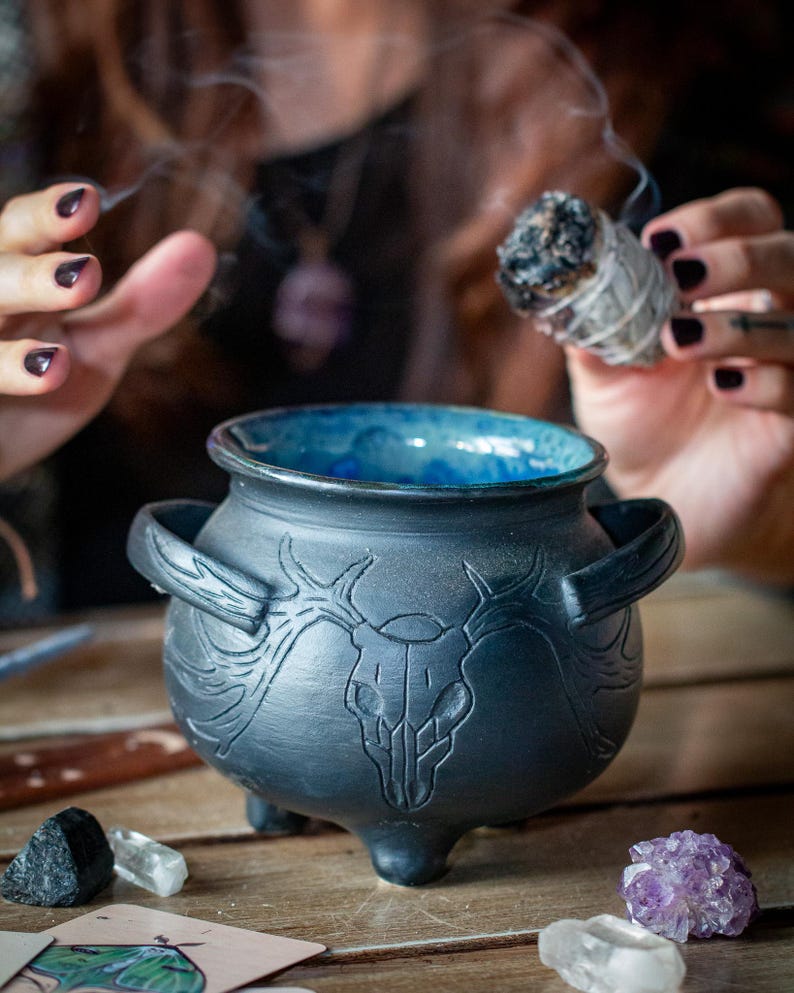 May include: A matte black ceramic cauldron featuring a carved deer skull design on the front, two side handles, and three small feet. The interior of the cauldron is glazed in a vibrant blue color, contrasting with the dark exterior finish.