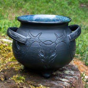 May include: A matte black ceramic cauldron featuring a relief carving of a deer skull with large antlers on the front. The vessel has two side handles, three small feet, and a glossy, deep blue glazed interior that contrasts with the dark exterior.