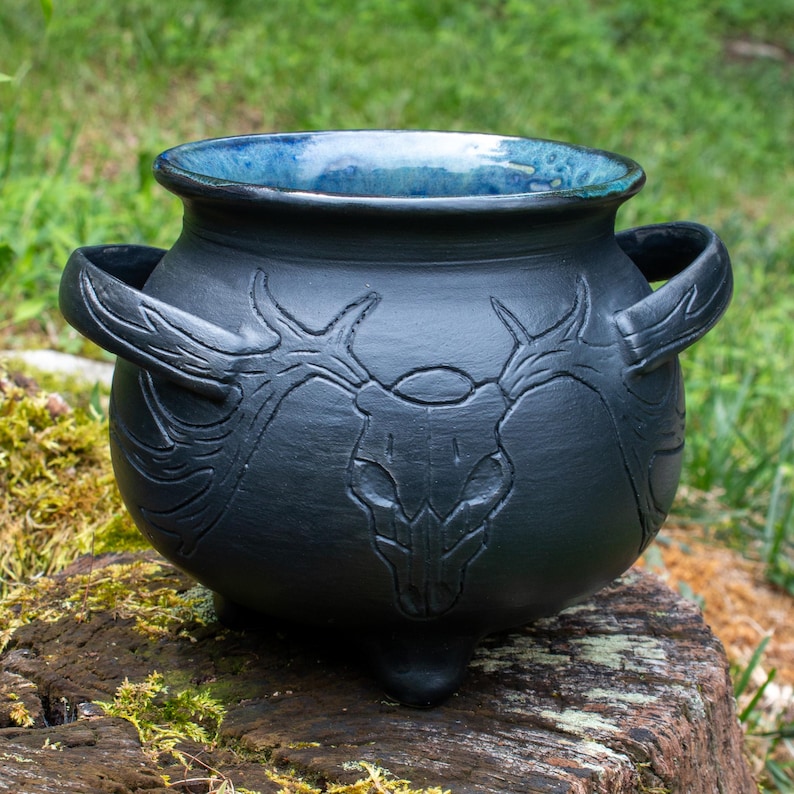 May include: A matte black ceramic cauldron featuring a relief carving of a deer skull with large antlers on the front. The vessel has two side handles, three small feet, and a glossy, deep blue glazed interior that contrasts with the dark exterior.