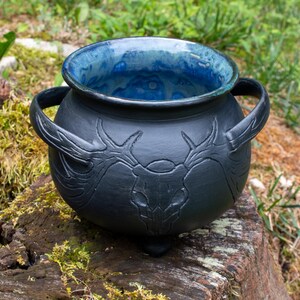 May include: A matte black ceramic cauldron with two side handles and three small feet. The exterior features a relief carving of a deer skull with large antlers. The interior is glazed in a vibrant, mottled blue finish with a smooth texture.