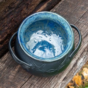 May include: A round ceramic pot with two side handles, featuring a matte black exterior with a textured, embossed pattern and a glossy, vibrant blue interior glaze that transitions into lighter shades of turquoise and white at the base.