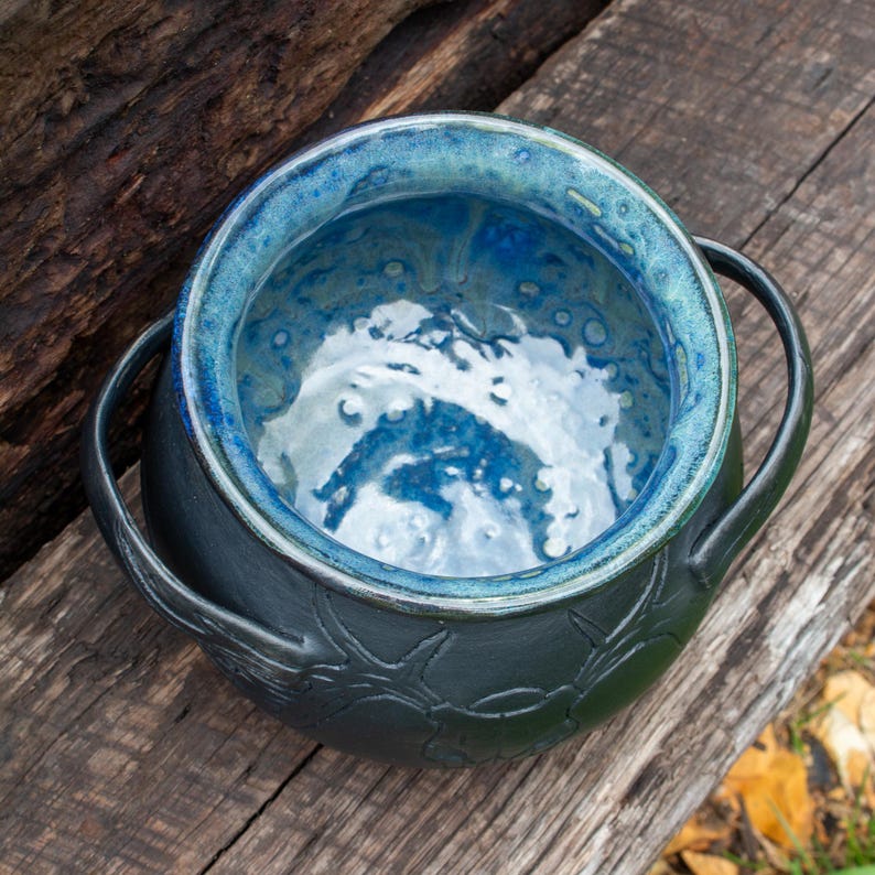 May include: A round ceramic pot with two side handles, featuring a matte black exterior with a textured, embossed pattern and a glossy, vibrant blue interior glaze that transitions into lighter shades of turquoise and white at the base.