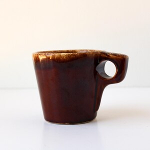 May include: A brown ceramic mug with a unique handle design. The mug has a dark brown exterior with a lighter brown rim. The handle features a circular opening. The mug is likely vintage.