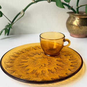 May include: Amber-colored glass teacup and saucer set. The saucer has a textured sunburst design. The teacup sits on the saucer, and both pieces have a warm, inviting hue. The set is ideal for serving tea or coffee.