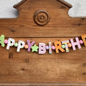 May include: A decorative garland spelling out the words HAPPY BIRTHDAY with individual wooden letters in various pastel colors, patterns, and shapes. The letters are strung together with small round beads and interspersed with star and flower accents.