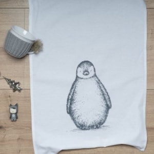 May include: White kitchen towel with a detailed illustration of a penguin. A gray mug with a knitted cozy and a small pom-pom is on the left. A small star-shaped ornament and a figurine are also visible.