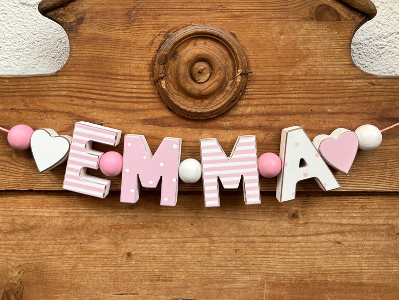 Może przedstawiać: Dekoracyjna girlanda z drewnianymi literami układającymi się w imię EMMA. Litery E i M mają r&oacute;żowo-białe paski, a litery M i A są r&oacute;żowo-białe w kropki. Girlanda zawiera r&oacute;żowe i białe koraliki oraz elementy w kształcie serc.