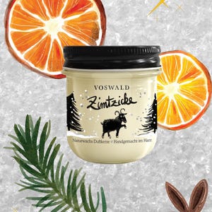 May include: A jar candle with a black lid, featuring the text "Zimtzicke" and a black goat illustration. The candle is surrounded by orange slices, a pine branch, and a star anise. The background is a textured, light gray.