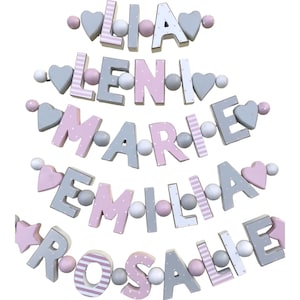 May include: A set of five wooden name garlands with pink and gray letters and beads. The names are Lia, Leni, Marie, Emilia, and Rosalie.