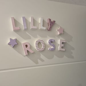 Może przedstawiać: Zestaw tr&oacute;jwymiarowych drewnianych liter tworzących napis LILLY ROSE w pastelowych odcieniach r&oacute;żu i fioletu. Litery posiadają r&oacute;żne wzory, w tym paski i kropki, a całość uzupełniają dwie pięcioramienne gwiazdki.