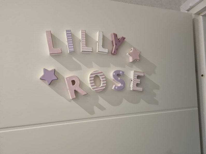 Może przedstawiać: Zestaw tr&oacute;jwymiarowych drewnianych liter tworzących napis LILLY ROSE w pastelowych odcieniach r&oacute;żu i fioletu. Litery posiadają r&oacute;żne wzory, w tym paski i kropki, a całość uzupełniają dwie pięcioramienne gwiazdki.
