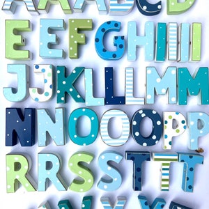 May include: A full alphabet of wooden letters in various shades of blue and green. Each letter is hand-painted with stripes, polka dots, or solid colors. The letters are arranged in rows, showcasing the entire alphabet.