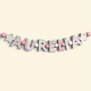 May include: A decorative garland spelling the name AURELIA with individual wooden letters. The letters alternate between light pink stripes and solid white with polka dots. The string is accented with pink and white wooden beads and stars.