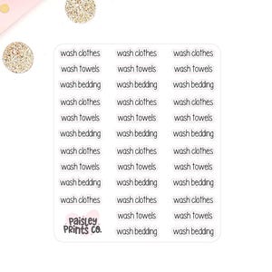 May include: A sheet of white stickers with black text, including phrases like "wash clothes," "wash towels," and "wash bedding." The stickers are arranged in rows and columns. The "Paisley Prints Co." logo is visible. Decorative pink flowers and gold glitter circles are in the background.
