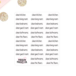 May include: A collection of white rectangular stickers with black text, including phrases like "clean kitchen," "clean living room," and "clean bedrooms." The stickers are arranged on a pink surface with gold glitter accents and pink flowers.