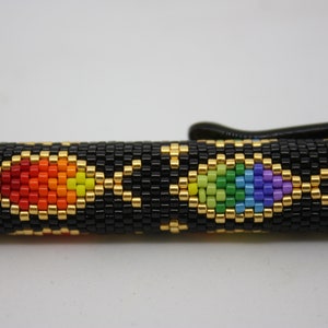 May include: Black pen with a beaded sleeve. The sleeve is adorned with a pattern of black, gold, and rainbow-colored beads. The rainbow colors include red, orange, yellow, green, blue, and purple.