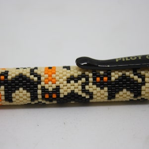 May include: A black pen with a beige and black beaded design featuring orange accents. The pen has the text "PILOT G-2 0.7" on the top.