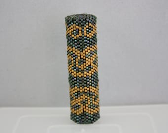 May include: A beaded cylinder with a geometric pattern in shades of gold and green.