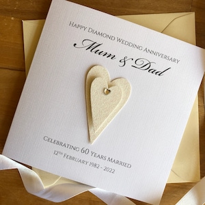 May include: A white card with the text "Happy Diamond Wedding Anniversary Mum & Dad Celebrating 60 Years Married 12th February 1982 - 2022" and two white layered hearts with a rhinestone in the centre.