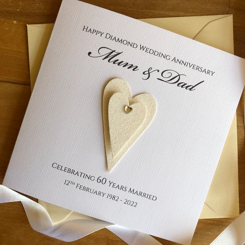 May include: A white card with the text "Happy Diamond Wedding Anniversary Mum & Dad Celebrating 60 Years Married 12th February 1982 - 2022" and two white layered hearts with a rhinestone in the centre.