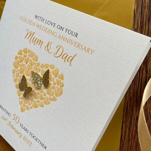May include: A white card with gold lettering that says "Golden Wedding Anniversary" and "With Love On Your Mum & Dad". The card has a gold heart shape made of smaller hearts and two gold glitter butterflies. The text at the bottom of the card says "Celebrating 50 Years Together 24th February 2023".