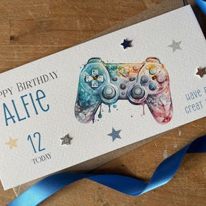 May include: A birthday card featuring a watercolour video game controller. The card says "Happy Birthday Alfie 12 Today" with star decorations and "Have a great day". A blue ribbon is in the foreground.