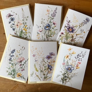 May include: Six greeting cards with watercolour floral designs. Each card displays a unique arrangement of wildflowers in shades of blue, purple, pink, and yellow, set against a textured white background. The cards have a pale yellow border.