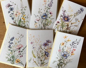 May include: A collection of six greeting cards with floral watercolor designs. Each card features a different arrangement of wildflowers in shades of blue, purple, pink, and yellow, set against a textured white background. The cards have a light yellow border.