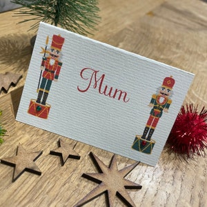 May include: A white place card with the word "Mum" printed in red. The card features a cartoon illustration of two nutcracker soldiers, one on each side of the text. The nutcracker soldiers are wearing red and gold uniforms and holding a weapon.