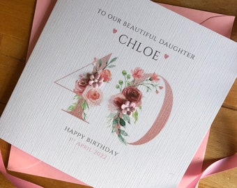 Puede incluir: Tarjeta de felicitación blanca para un 40 cumpleaños, con el nombre "Chloe" y el texto "A nuestra hermosa hija". El número 40 es de color oro rosa y está decorado con arreglos florales. La tarjeta está sobre un sobre rosa.